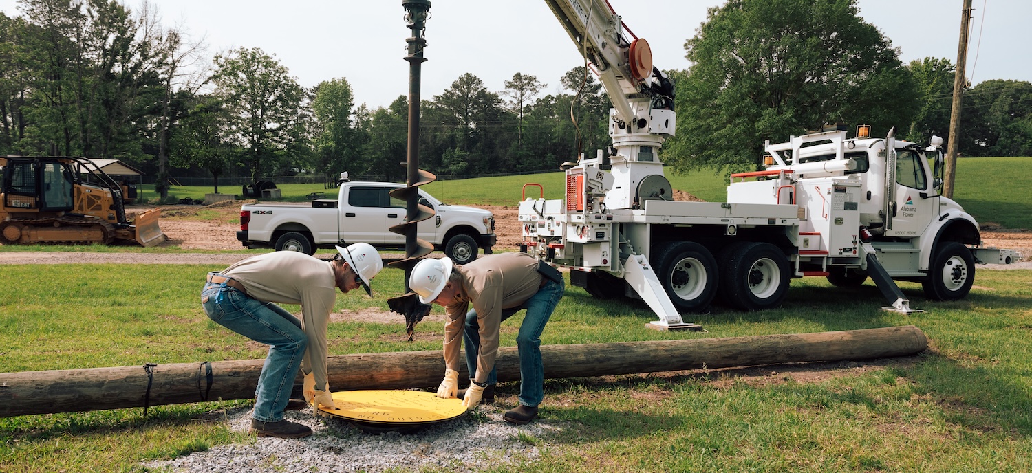Image of two lineman placing a yellow, round cover over a hole dug by an Altec digger derrick.